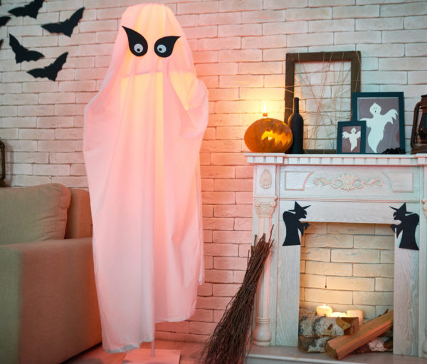 Common living room with sofa and fireplace decorated with Halloween toys and crafts such as lanterns, frames with ghosts, paper bats and withes, big ghost from fabric and broom